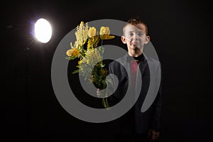 a boy with vitiligo in a red T-shirt, gray jacket and bow tie, with a bouquet of mimosa and yellow tulips