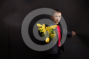 a boy with vitiligo in a red T-shirt, gray jacket and bow tie, with a bouquet of mimosa and yellow tulips