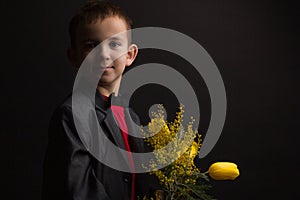 a boy with vitiligo in a red T-shirt, gray jacket and bow tie, with a bouquet of mimosa and yellow tulips