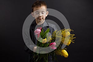 a boy with vitiligo in a red T-shirt, gray jacket and bow tie, with a bouquet of mimosa and yellow tulips