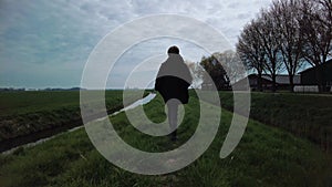 Boy walking alone on a dike with green grass in the Dutch countryside, rear view