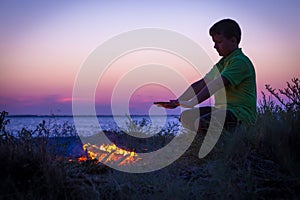 Boy warms by the campfire on the beach at sunset