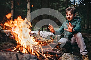 Boy warms his hands near campfire in forest