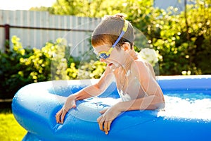 A boy in water glasses swims in an inflatable pool in the garden on a sunny summer day