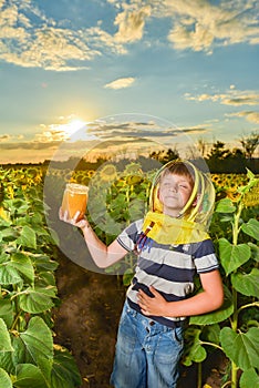 A boy wearing a beekeeper mask holds a jar of honey in the middle of a sunflower field and imagines how he eats honey