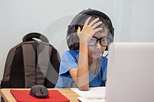 A boy wearing headphones and light filtering glasses is using laptops computers and puts his hands over his head due to the st
