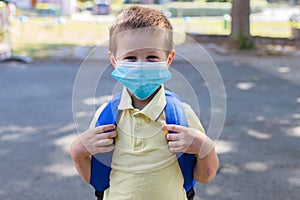 Boy wearing a protective mask with a backpack behind his back in the schoolyard on the first school day after isolation or