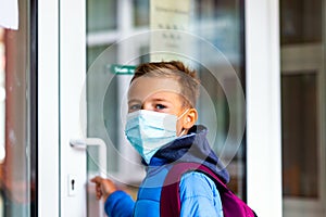 The boy wearing protective mask is trying to open the school door. Behind the backpack Schoolboy look at camera