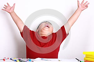 The boy wearing a red shirt happily stretched out his arm and sat on the desk.