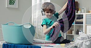 Boy wearing wireless headphones stands on stool in laundry room by fold-out dryer. A child is listening to music and