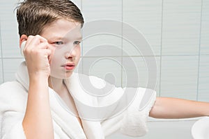 A boy in a white bathrobe in the bathroom holds soap in his ear like a phone