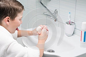 A boy in a white bathrobe in the bathroom washes his hands with a bar of soap in the sink