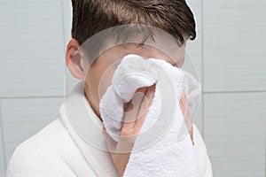 Boy, in a white bathrobe, in the bathroom, wipes his face with a towel, after washing