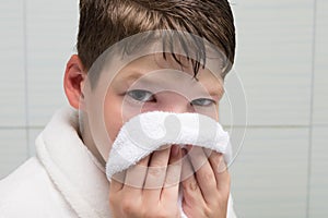 Boy, in a white bathrobe, in the bathroom, wipes his face with a towel, after washing, close-up