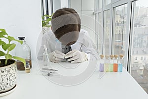 Boy in white gloves is looking into little microscope, five colorful test tubes stand near on table,