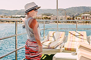 A boy in a white hat and shorts holding the railing on the ship and looking at the sea