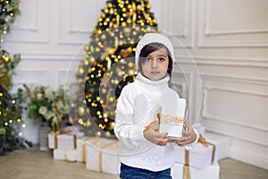 boy in a white knitted sweater and hat stands with a gift box at the Christmas tree at home on Christmas Day