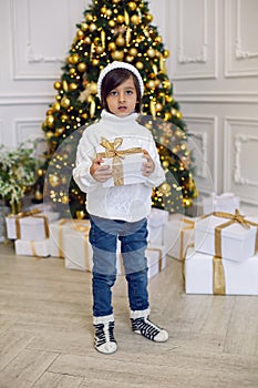 boy in a white knitted sweater and hat stands with a gift box at the Christmas tree at home on Christmas Day