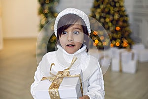 boy in a white knitted sweater and hat stands with a gift box at the Christmas tree at home on Christmas Day