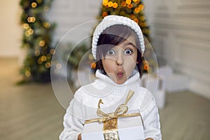 boy in a white knitted sweater and hat stands with a gift box at the Christmas tree at home on Christmas Day