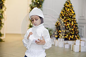 boy in a white knitted sweater and hat stands with a gift box at the Christmas tree at home on Christmas Day