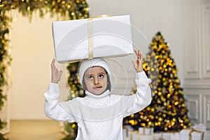 boy in a white knitted sweater and hat stands with a gift box at the Christmas tree at home on Christmas Day