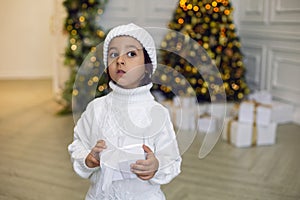 boy in a white knitted sweater and hat stands with a gift box at the Christmas tree at home on Christmas Day