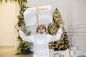 boy in a white knitted sweater and hat stands with a gift box at the Christmas tree at home on Christmas Day