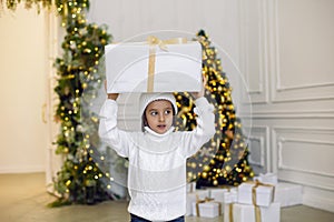 boy in a white knitted sweater and hat stands with a gift box at the Christmas tree at home on Christmas Day