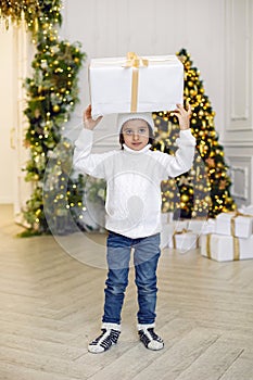 boy in a white knitted sweater and hat stands with a gift box at the Christmas tree at home on Christmas Day
