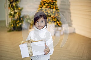 boy in a white knitted sweater stands with a gift box at the Christmas tree at home on Christmas Day