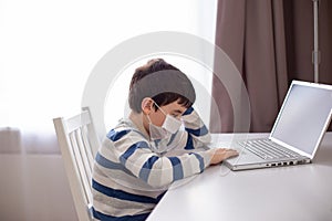 A boy in a white medical mask , sits behind a laptop