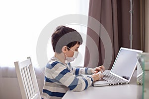 A boy in a white medical mask , sits behind a monitor with a laptop in room