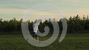 A boy in a white shirt running through a summer sunny field with a toy plane in his hand above his head. The boy