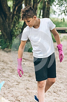 Boy in white t shirt in gloves collects garbage and plastic bottles into blue package on the beach.