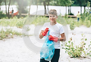 Boy in white t shirt in gloves collects garbage and plastic bottles into blue package on the beach. Young volunteer.