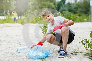 Boy in white t shirt in gloves collects garbage and plastic bottles into blue package on the beach. Young volunteer.