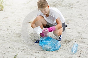 Boy in white t shirt in gloves collects garbage and plastic bottles into blue package on the beach. Young volunteer