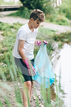 Boy in white t shirt in gloves collects garbage and plastic bottles into blue package on the beach. Young volunteer