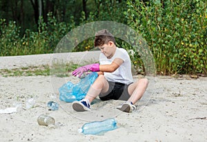 Boy in white t shirt in gloves collects garbage and plastic bottles into blue package on the beach. Young volunteer