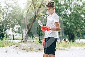 Boy in white t shirt in gloves collects garbage and plastic bottles into blue package on the beach. Young volunteer