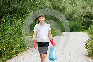 Boy in white t shirt in gloves collects garbage and plastic bottles into blue package on the beach. Young volunteer