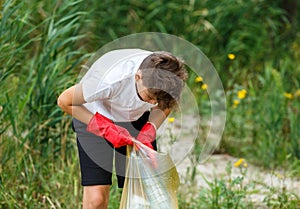 Boy in white t shirt in gloves collects garbage and plastic bottles into blue package on the beach. Young volunteer. Environment