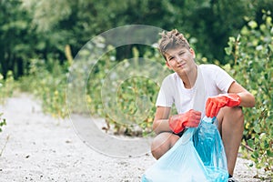 Boy in white t shirt in gloves collects garbage and plastic bottles into blue package on the beach. Young volunteer. Environment