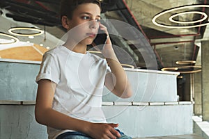Boy in white t-shirt is sitting indoors and talking on his mobile phone. A teenager uses a cell phone, calling, phoning