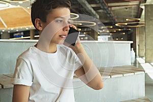 Boy in white t-shirt is sitting indoors and talking on his mobile phone. A teenager uses a cell phone, calling, phoning