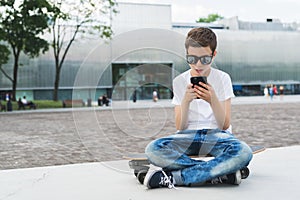Boy in white T-shirt and sunglasses sitting outdoor on longboard and uses smartphone. Boy plays computer games