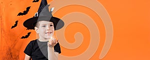 A boy in a witch costume in a black hat holds a small flashlight glowing pumpkin lamp Jack. on an orange background