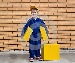 Boy of 6 years old in a blue overalls - uniform stands near a brick wall, holds yellow cardboard parcel boxes photo