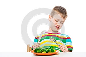 A boy of 10 years old looks at broccoli in a plate in disgust, portrait is isolated on a white